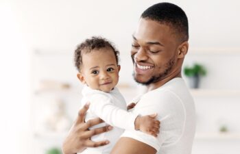 Happy African American Father Cuddling His Adorable Newborn Baby Son At Home, Loving Black Dad Spending Time With Cute Infant Child, Holding Him On Hands, Enjoying Paternity Leave, Copy Space